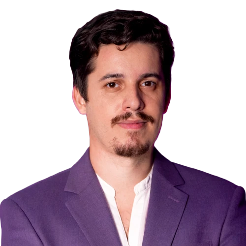 Foto de meio corpo do professor Guilherme Santos, da Escola de Pessoas. Ele tem cabelos escuros, bigode e cavanhaque, e veste um paletó roxo sobre uma camisa branca. O fundo é branco.